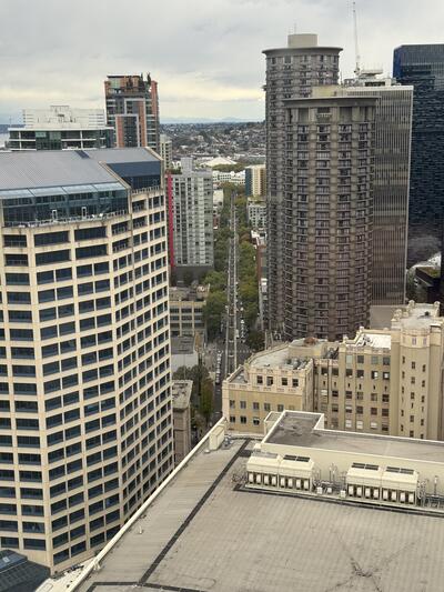 A cityscape photograph taken from a high vantage point showing downtown Seattle buildings including the cylindrical Westin Hotel, modern glass and older concrete buildings, with 5th avenue running vertically straight in the center of the image, under a cloudy sky.