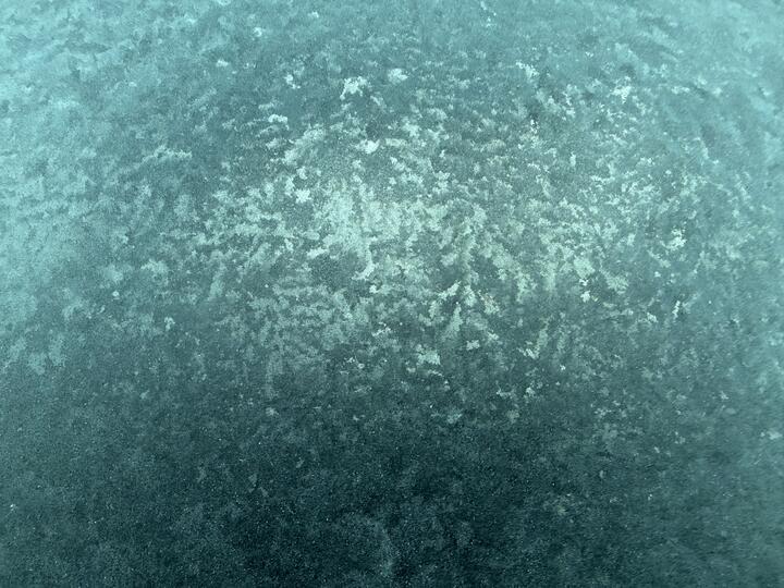 Close-up of frost on a car window, mottled teal and pale speckles.