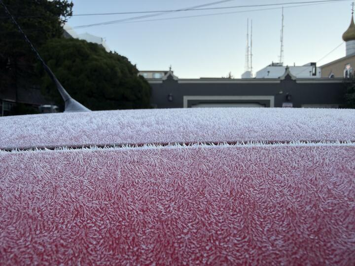 White frost crystals coat the roof of a red car, with a black antenna at left and a gray building and radio towers blurred in the background.