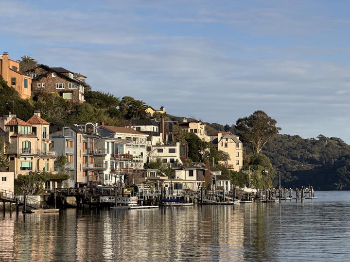 A row of houses and docks along a calm waterfront, with the buildings reflected in the water and low hills in the background.