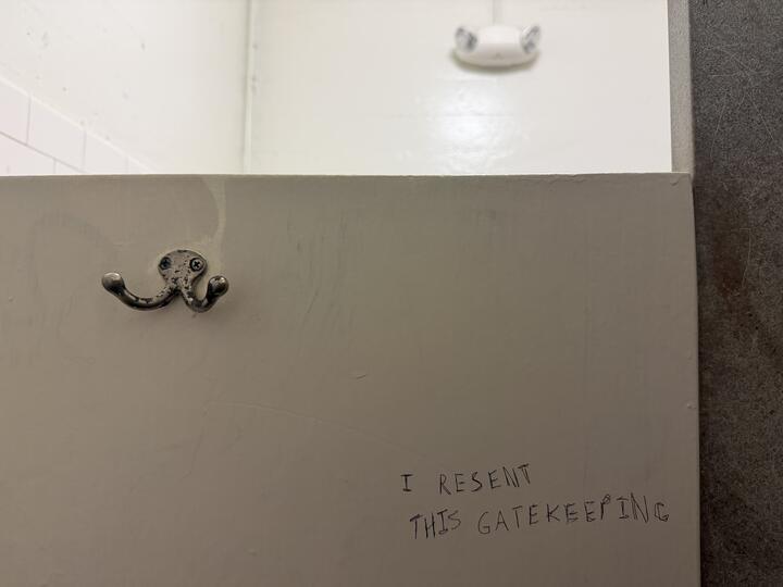 A photograph of a bathroom stall interior. A silver double-pronged coat hook is mounted on a cream-colored painted stall wall. Above the stall wall in the background, a small white motion sensor is visible on the white ceiling, a gray tile wall to the right. Near the bottom of the stall wall is graffiti that reads, in all caps, "I resent this gatekeeping."
