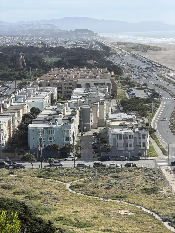 An elevated southern view of San Francisco's Outer Sunset neighborhood. Rows of pastel-colored residential buildings — soft blue, cream, and tan — stretch back in a grid toward the Pacific Ocean. The Great Highway runs along the coast lined with parked cars, with Ocean Beach and its sand dunes to the right. On the left edge, the historic Dutch windmill of Golden Gate Park is visible among trees. Hazy hills rise in the distance. A walking path between green coastal dunes fills the foreground.